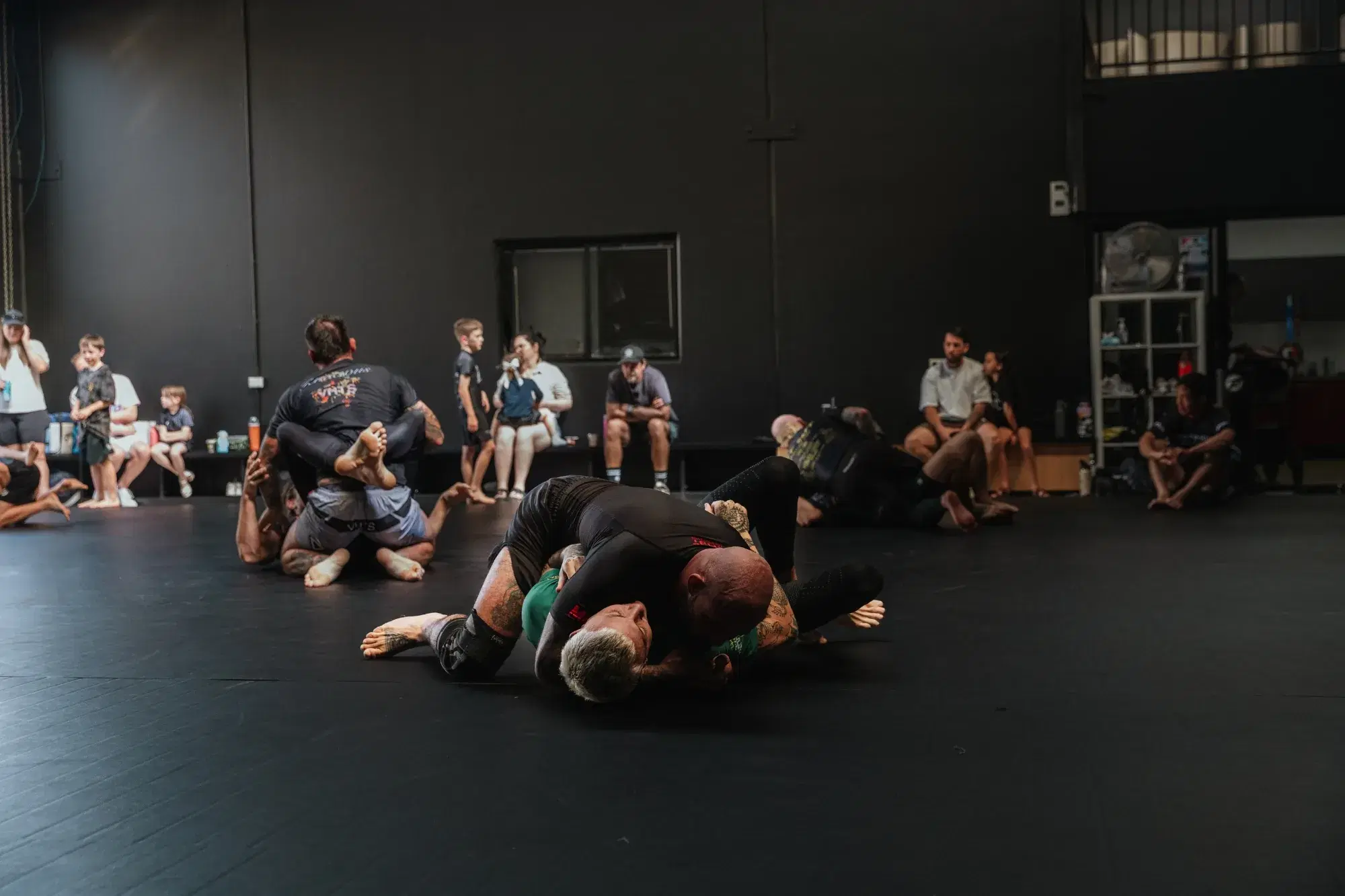 Adults training no-gi jiu-jitsu on the mats while families watch at Platinum Jiu Jitsu Oxenford